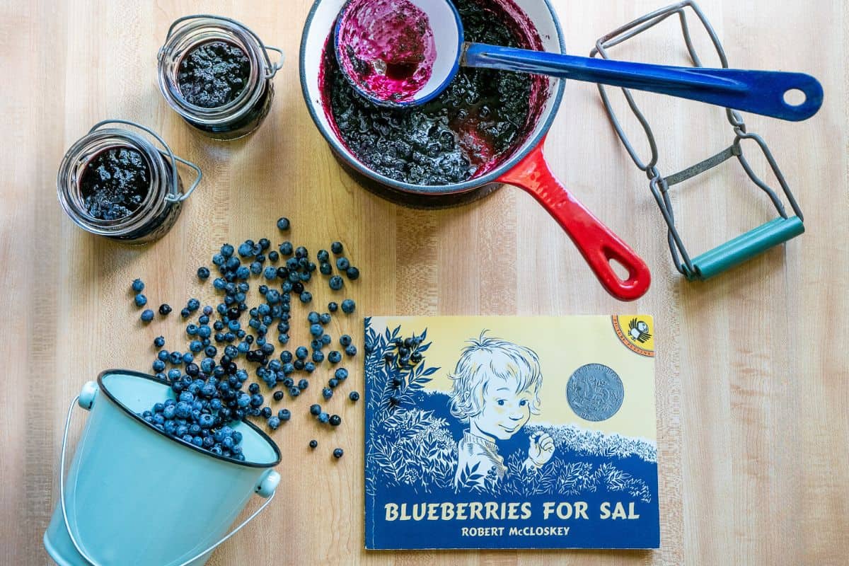 The cover of the picture book "Blueberries for Sal," surrounded by a bucket of blueberries spilling out onto the table, a pot of blueberry jam with a ladle, and two jars of blueberry jam.