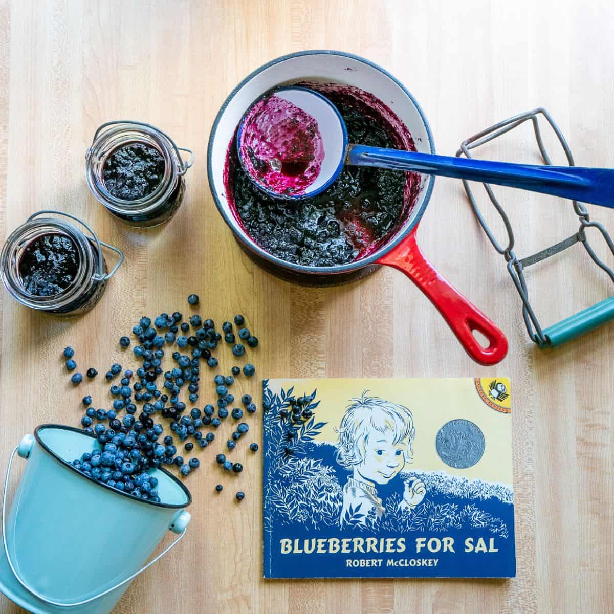 The cover of the picture book "Blueberries for Sal," surrounded by a bucket of blueberries spilling out onto the table, a pot of blueberry jam with a ladle, and two jars of blueberry jam.