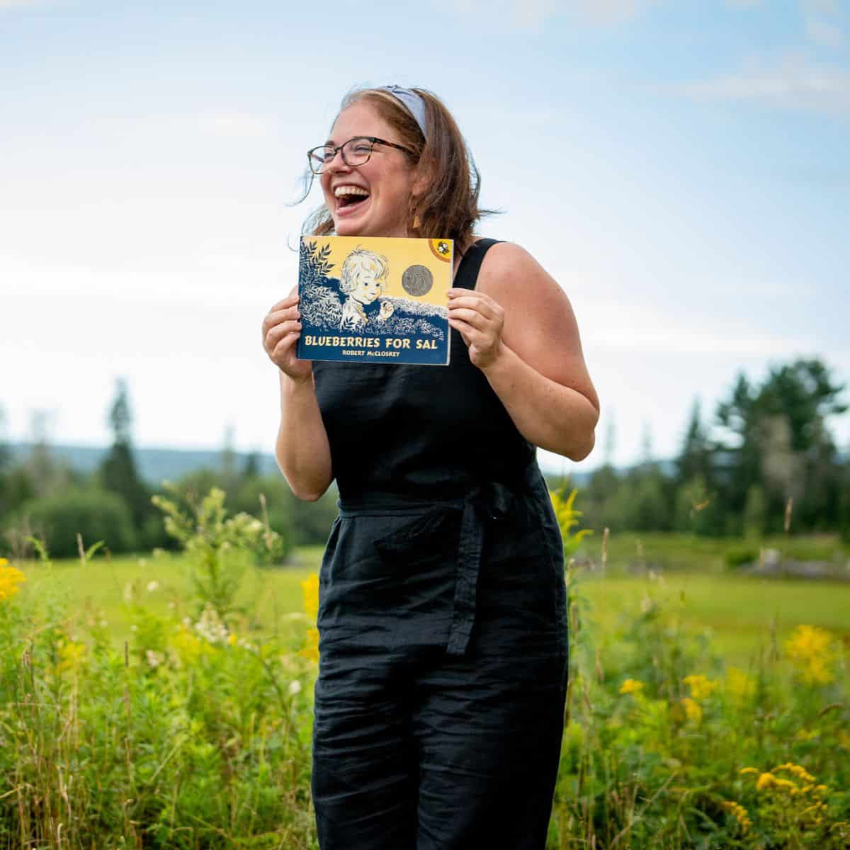 Afton Cyrus outdoors in a blueberry field, holding up the book Blueberries for Sal and laughing.