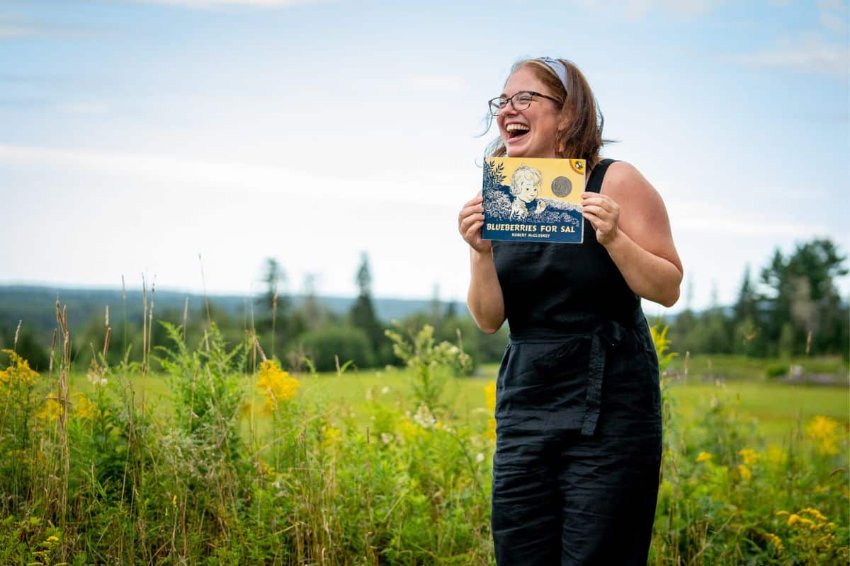 Afton Cyrus outdoors in a blueberry field, holding up the book Blueberries for Sal and laughing.