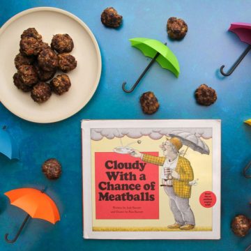 A plate of cooked oven-baked meatballs and the cover of the book "Cloudy with a Chance of Meatballs" surrounded by cooked meatballs and small colorful toy umbrellas.