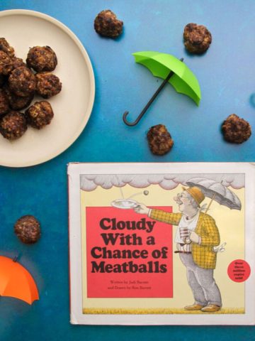 A plate of cooked oven-baked meatballs and the cover of the book "Cloudy with a Chance of Meatballs" surrounded by cooked meatballs and small colorful toy umbrellas.