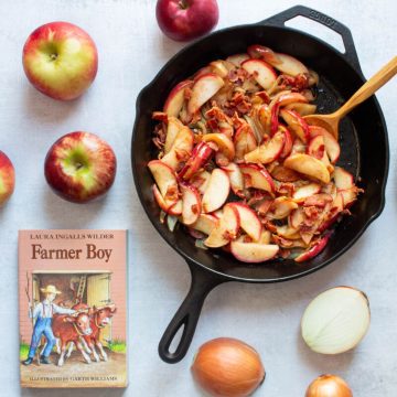 Fried Apples and Onions in a cast iron skillet, alongside the book "Farmer Boy" and whole apples and onions on a white surface.