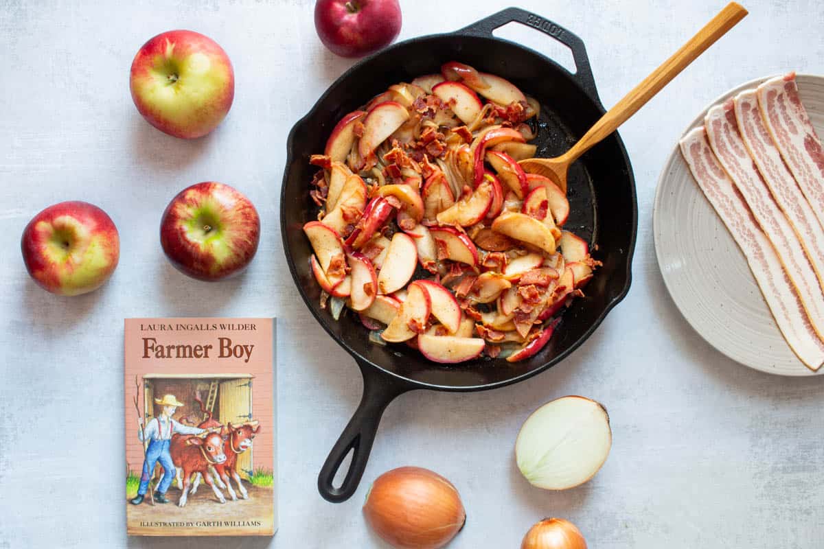 Fried Apples and Onions in a cast iron skillet, alongside the book "Farmer Boy" and whole apples and onions on a white surface.