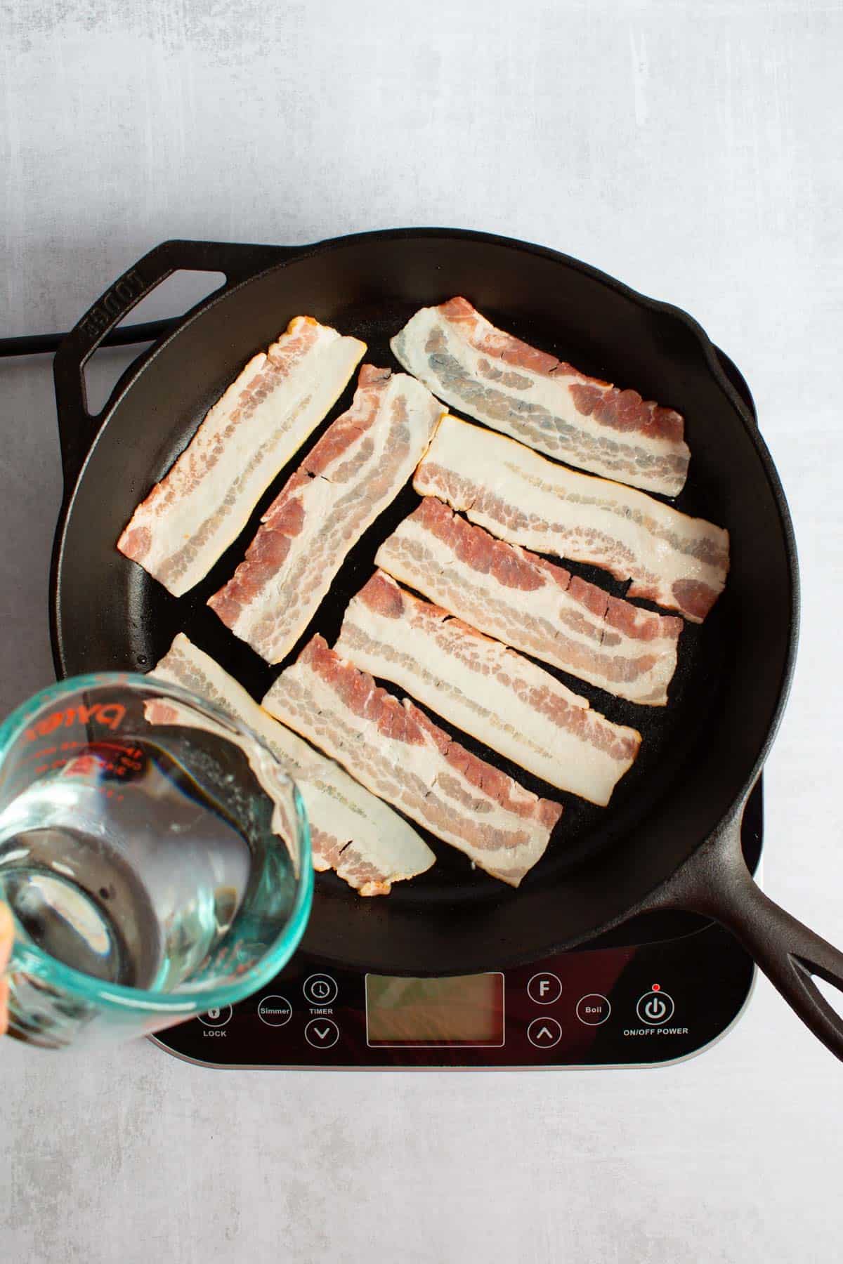 A liquid measuring cup pouring water into a cast iron skillet with slices of bacon in it for Apples and Onions.