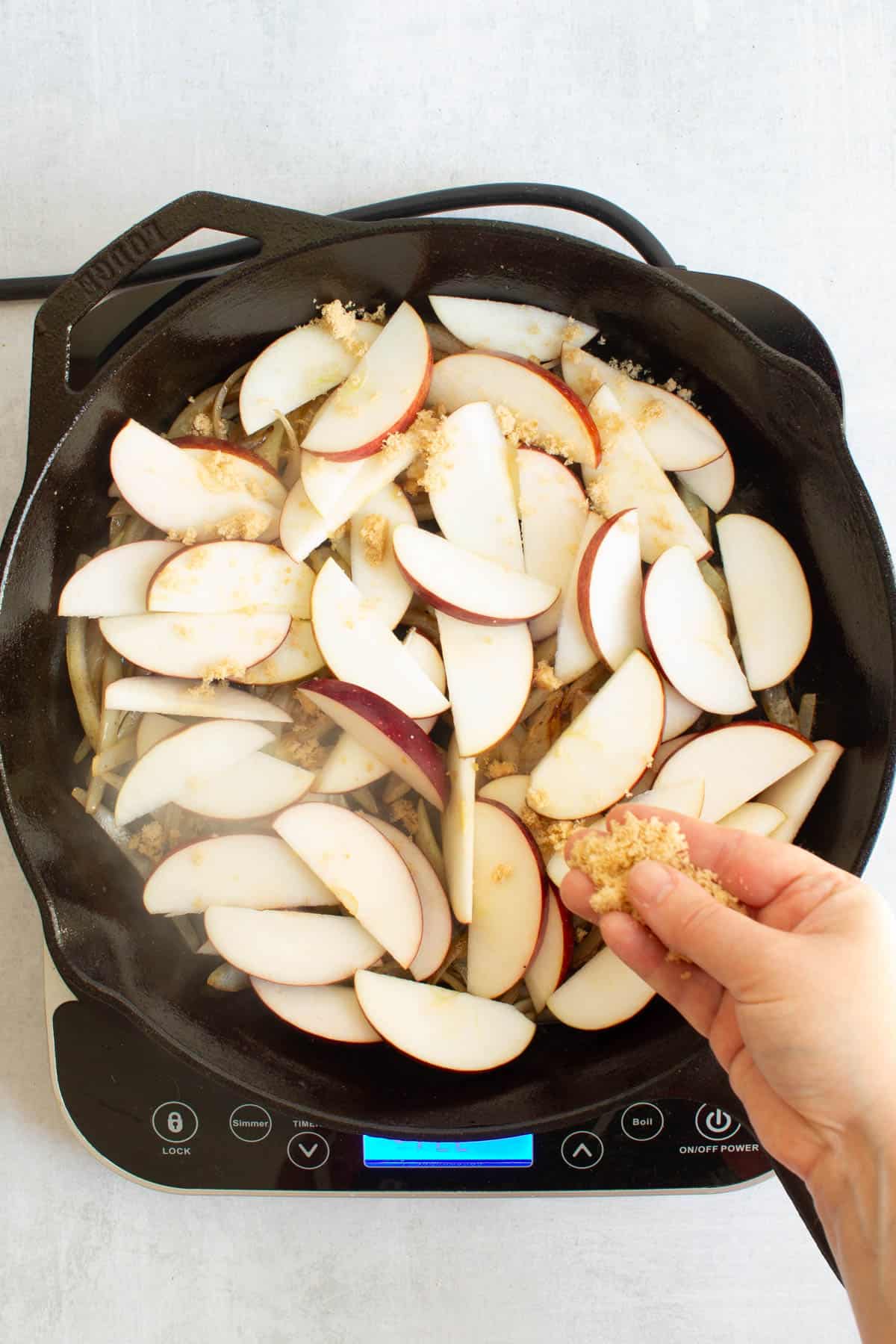 A hand sprinkling brown sugar over sliced apples in a skillet for Apples and Onions.