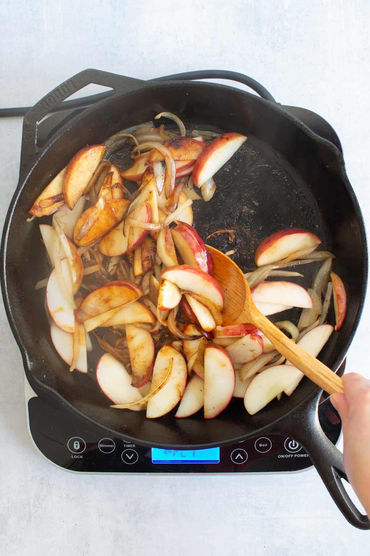 A hand using a wooden spoon to stir together cooked Apples and Onion in a cast iron skillet.