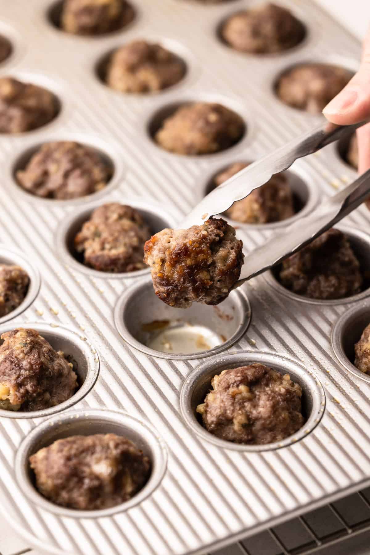 Tongs flipping a partially-baked meatball in a mini muffin tin.