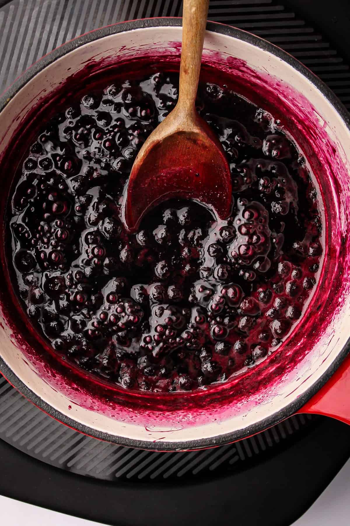 A wooden spoon stirring a batch of cooked wild Maine blueberry jam in a saucepan.