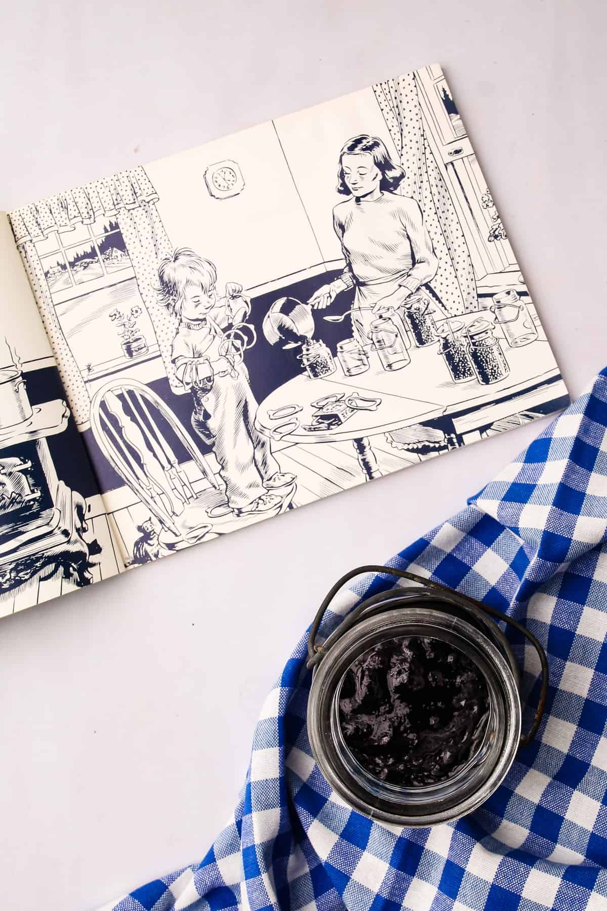 A jar of cooked wild Maine blueberry jam and the book "Blueberries for Sal" open to a page of illustration.