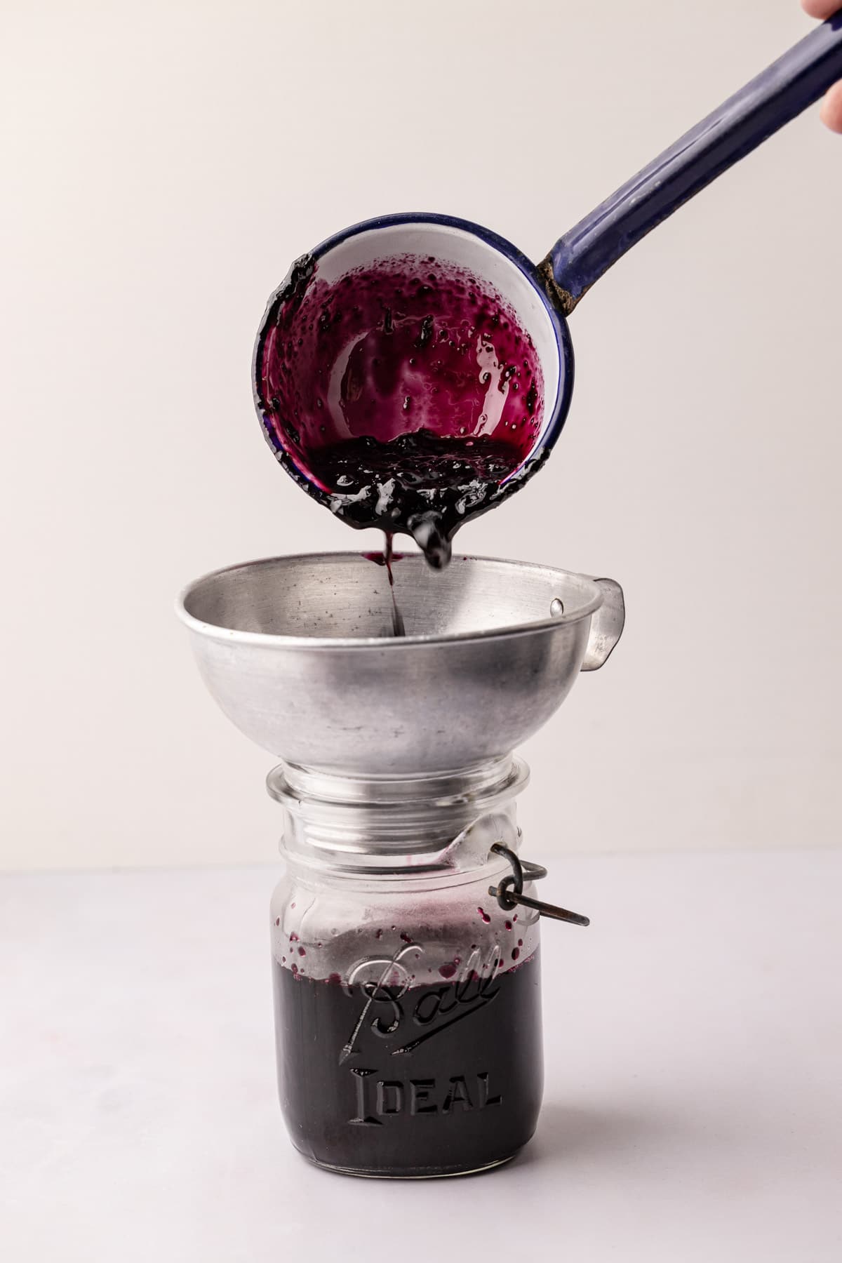 A ladle pouring wild Maine blueberry jam into a glass jar with a canning funnel in it.