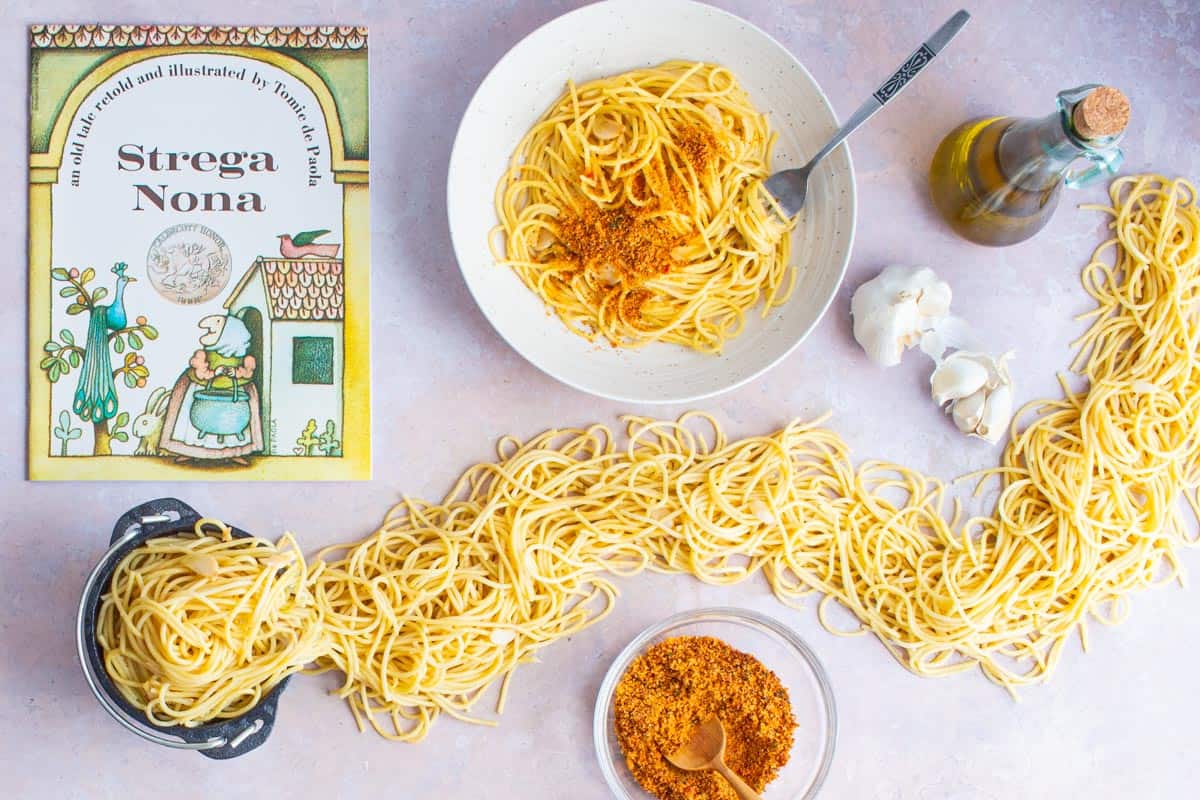 A bowl of Spaghetti and Garlic and Oil and a bowl of spicy Calabrian breadcrumbs, next to the book Strega Nona and a small cauldron with spaghetti spilling out of it all over the table.