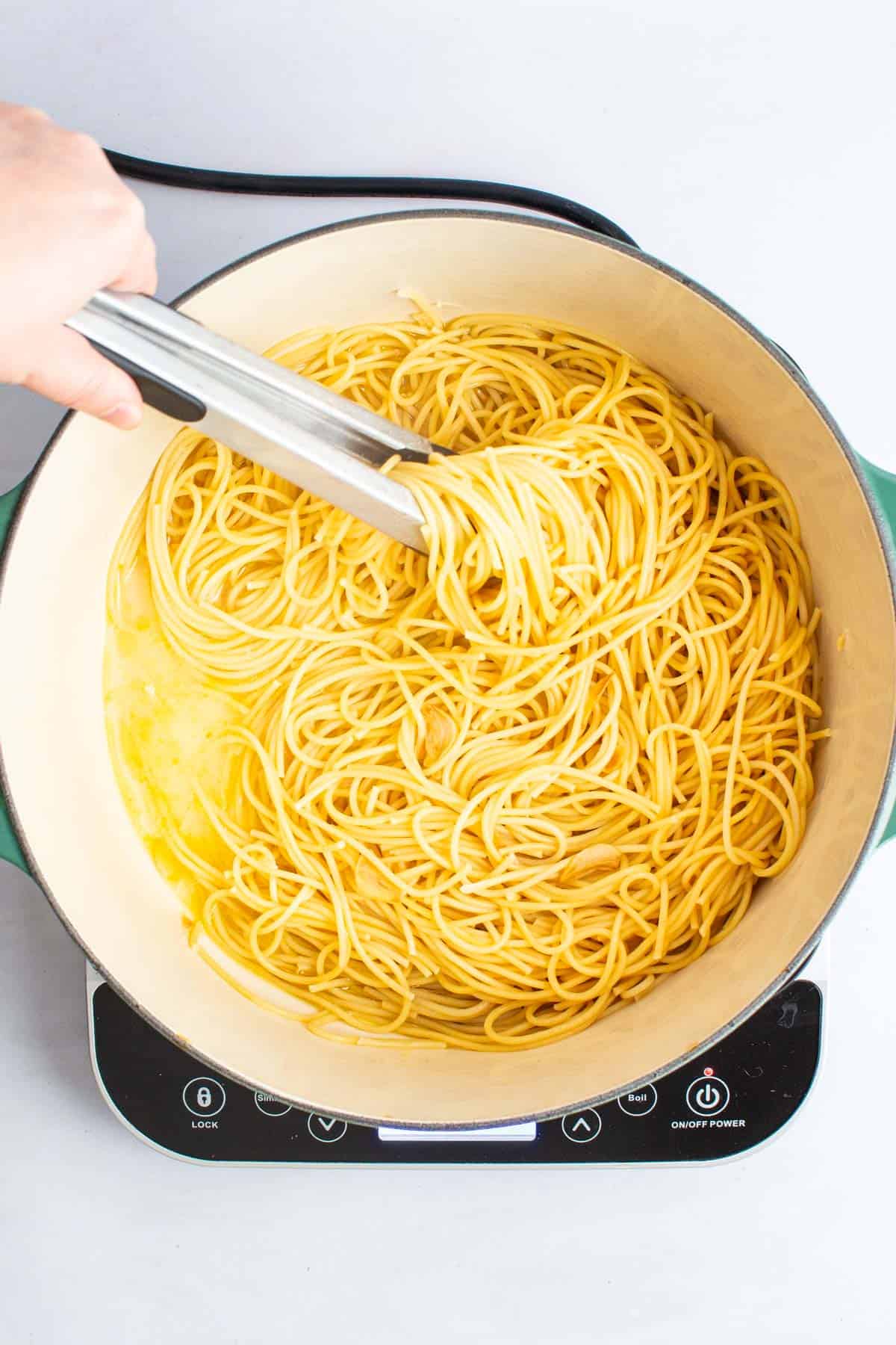 A hand using tongs to stir pasta in a pot for Spaghetti with Garlic and Oil.