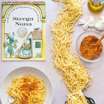 A bowl of Spaghetti and Garlic and Oil and a bowl of spicy Calabrian breadcrumbs, next to the book Strega Nona and a small cauldron with spaghetti spilling out of it all over the table.