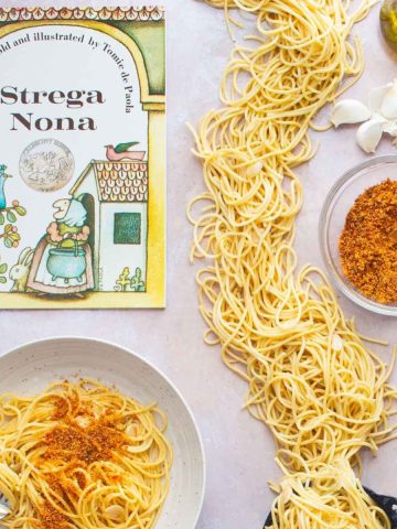 A bowl of Spaghetti and Garlic and Oil and a bowl of spicy Calabrian breadcrumbs, next to the book Strega Nona and a small cauldron with spaghetti spilling out of it all over the table.