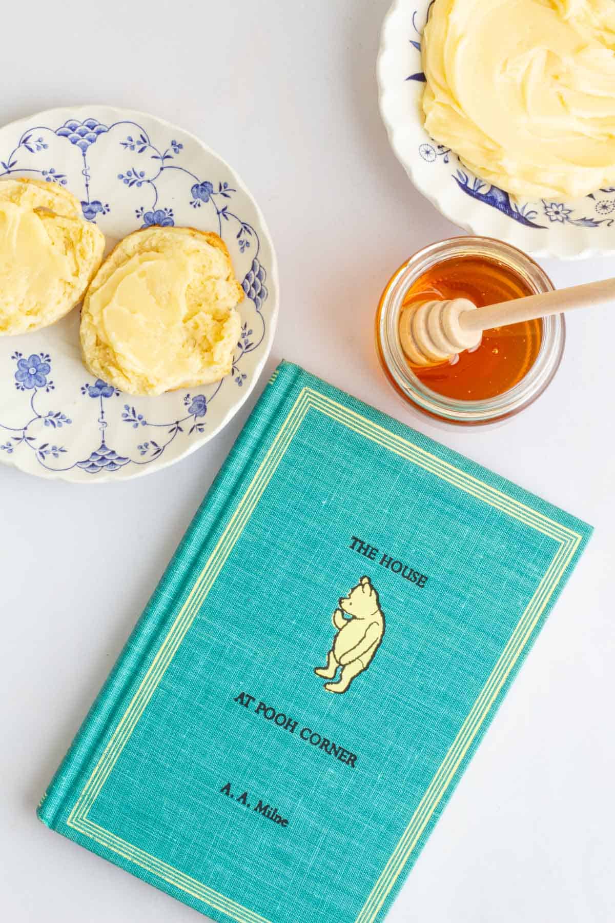 The cover of the book The House at Pooh Corner next to a plate of cream scones with homemade honey butter.