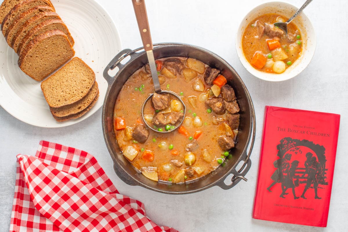 A pot and bowl of Dutch oven beef stew with a loaf of sliced bread, a red check napkin, and the book "The Boxcar Children" next to them.