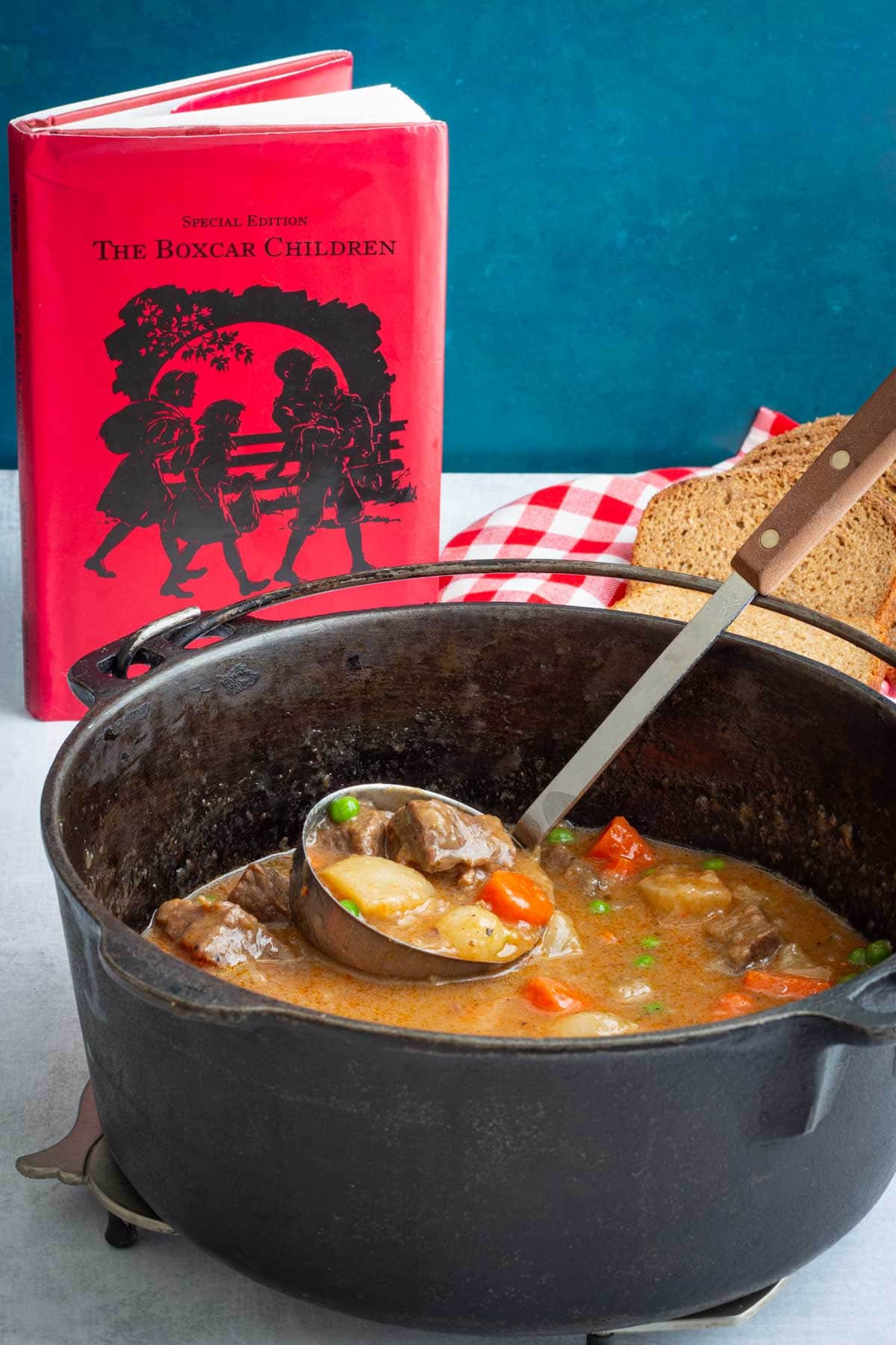 A pot and ladle filled with Dutch Oven Beef Stew, with the book The Boxcar Children and a loaf of bread next to it.