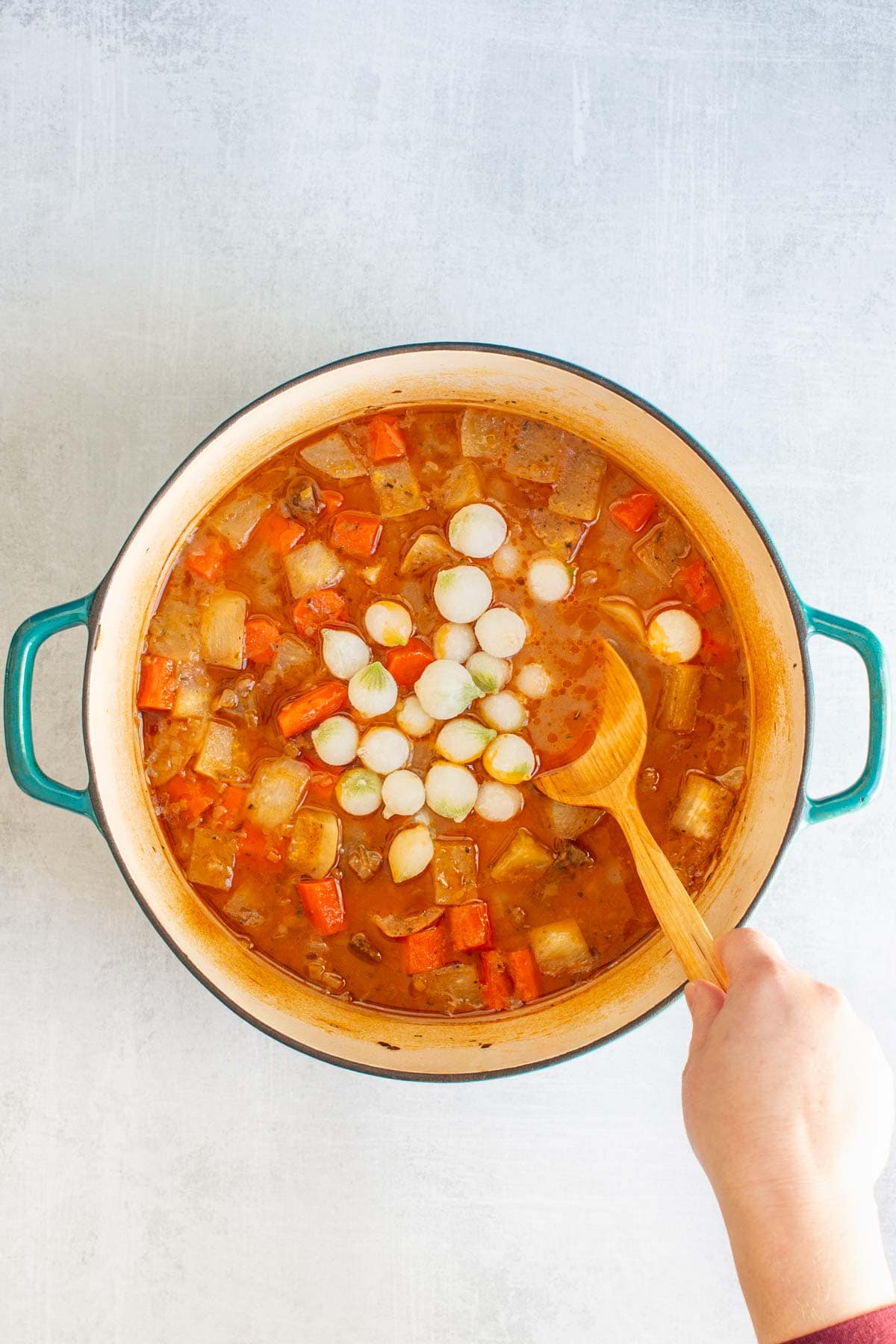 A hand using a wooden spoon to stir pearl onions into a pot of Dutch Oven Beef Stew.