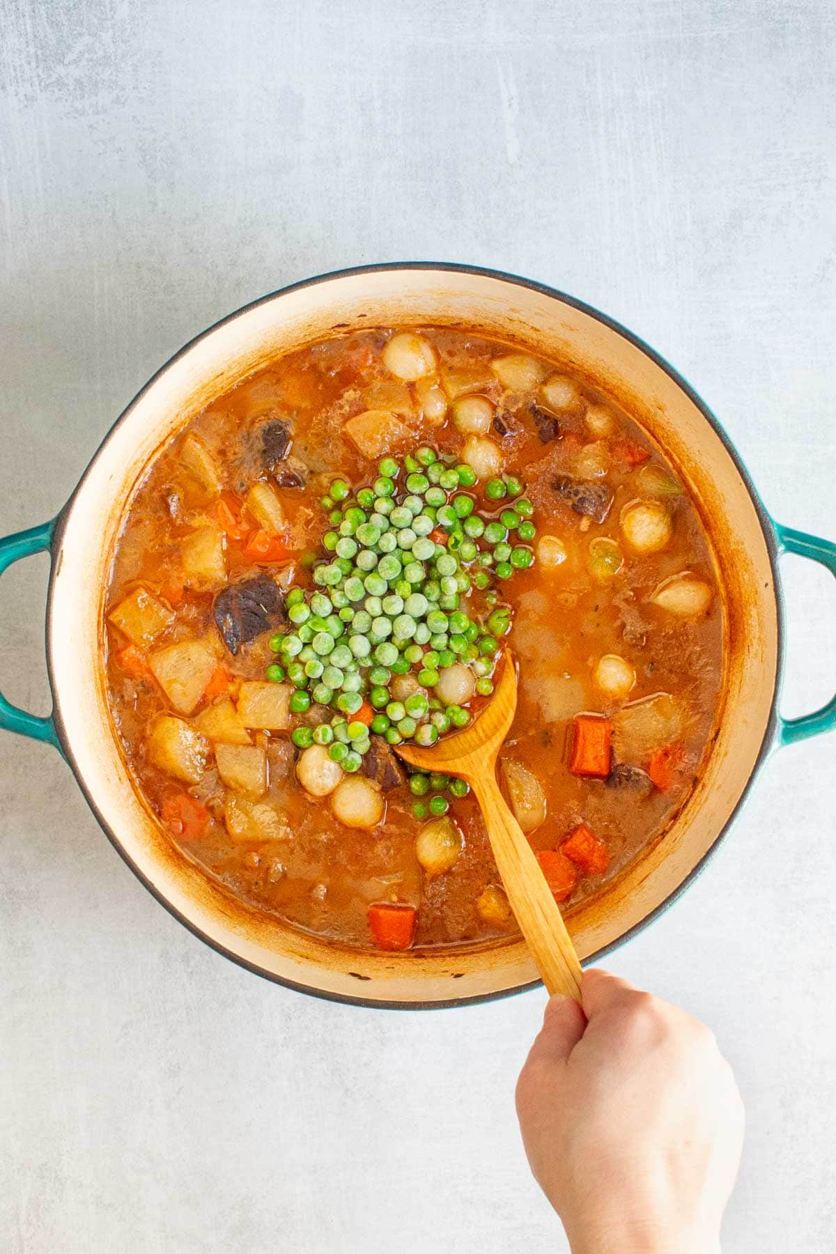 A hand using a wooden spoon to stir frozen peas into a pot of Dutch Oven Beef Stew.