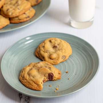 A plate with two small-batch chocolate chip cookies on it, one with a bite taken out, with a glass of milk and a plate of more cookies in the background.