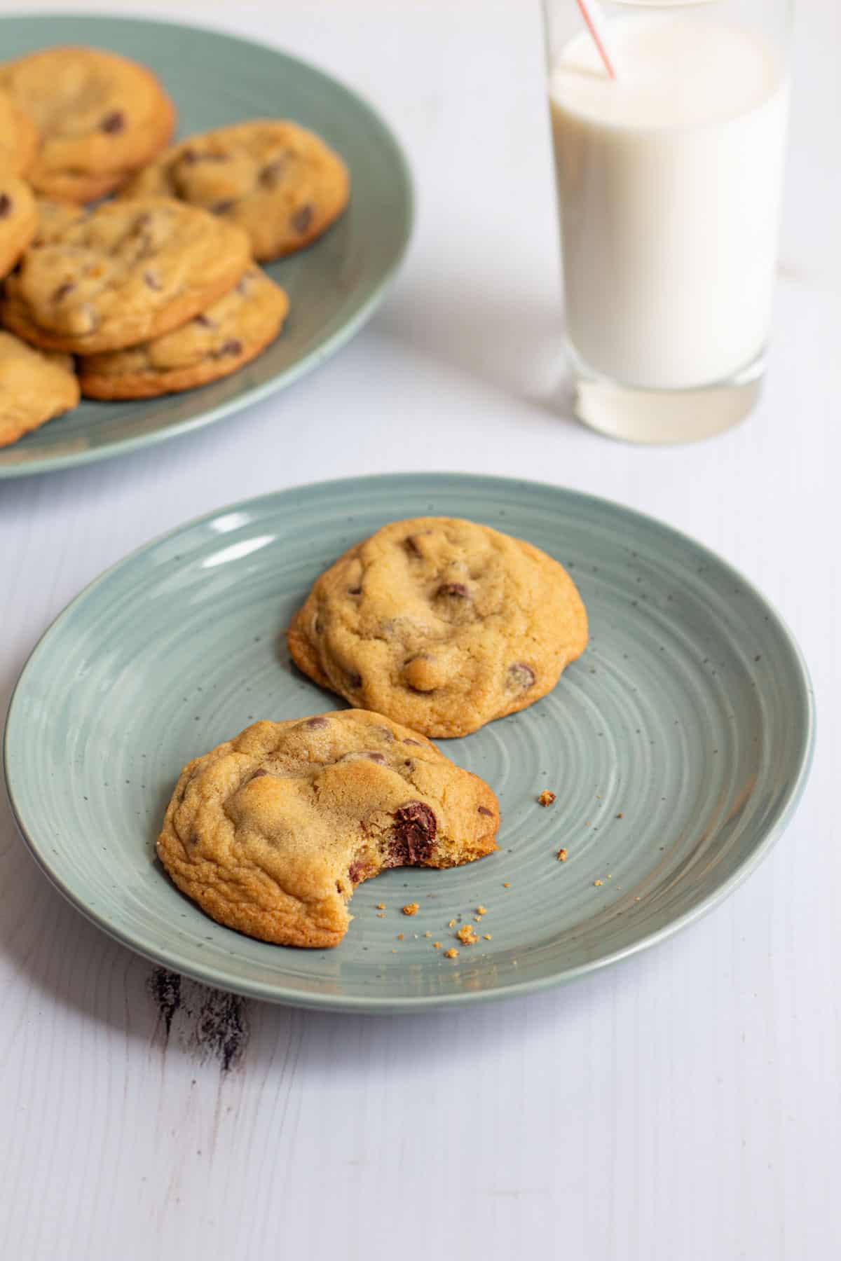 A plate with two small-batch chocolate chip cookies on it, one with a bite taken out, with a glass of milk and a plate of more cookies in the background.