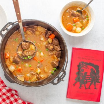 A pot and bowl of Dutch oven beef stew with a red check napkin and the book "The Boxcar Children" next to them.