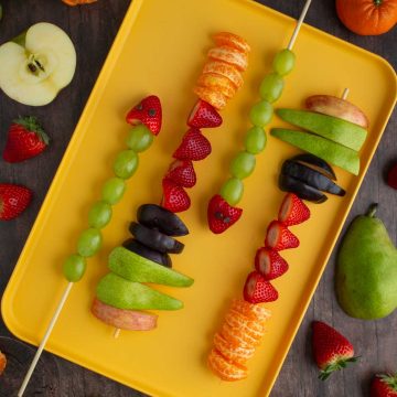 Assembled fruit skewers on a tray, with different fruits scattered on the surface nearby.