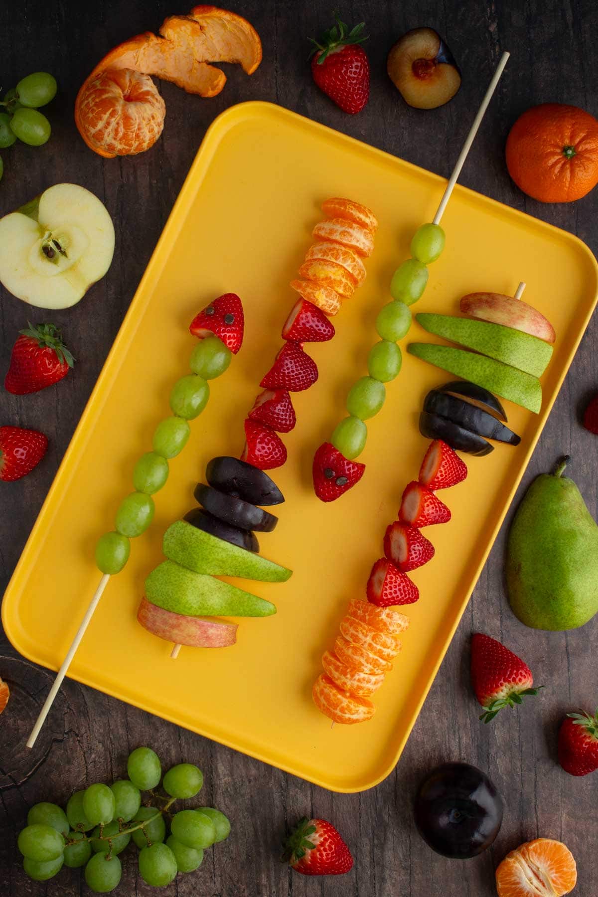 Assembled fruit skewers on a tray, with different fruits scattered on the surface nearby.