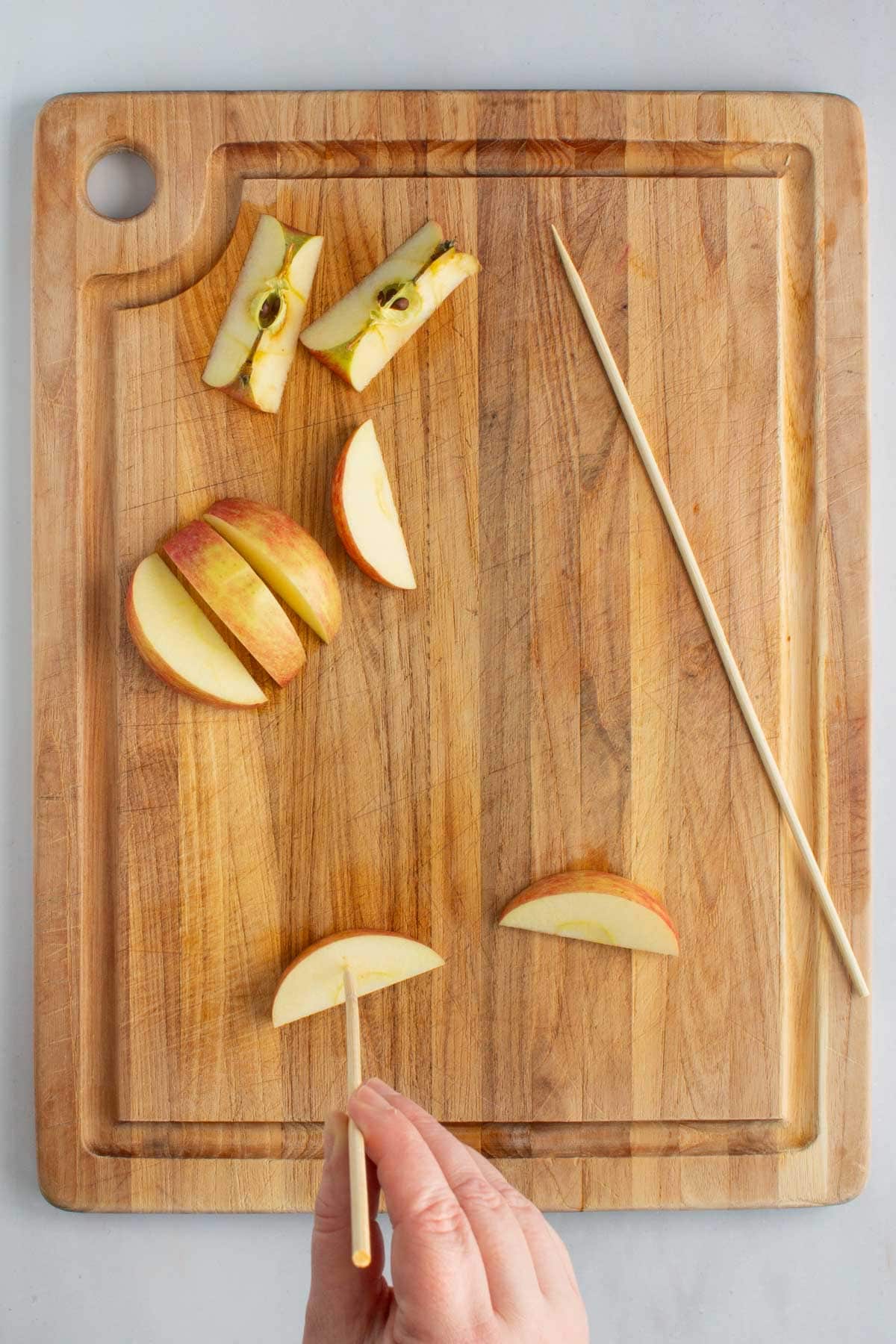 A hand poking an apple slice with a wooden skewer on a cutting board for fruit skewers, with more apple slices and another skewer nearby.