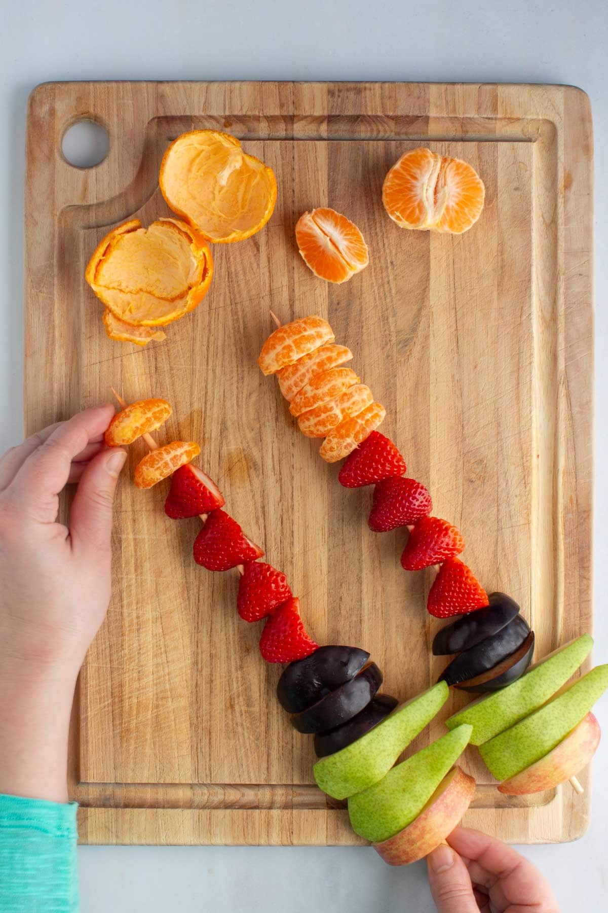 A hand sliding a clementine slice onto assembled fruit skewers on a cutting board.