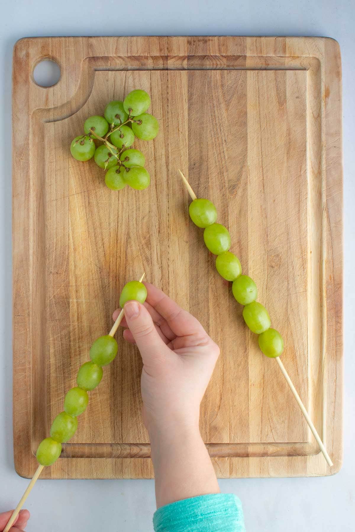 A hand sliding a green grape onto a wooden skewer for fruit skewers.