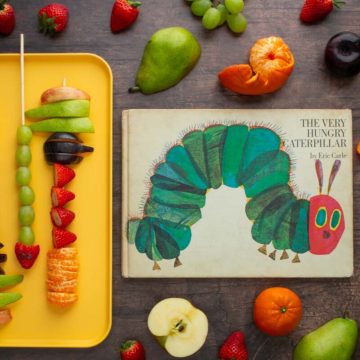 Assembled fruit skewers next to the cover of the book The Very Hungry Caterpillar, with different fruits scattered on the surface nearby.