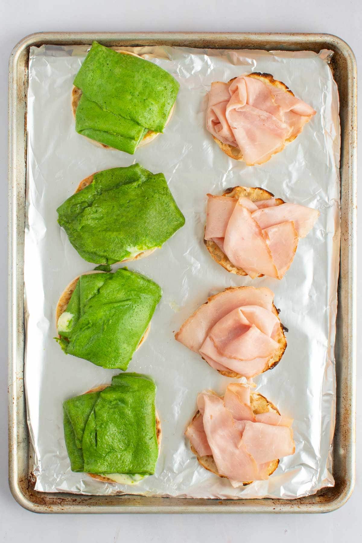 A baking sheet with four sets of cooked eggs and deli ham on English muffins for green eggs and ham breakfast sandwiches.