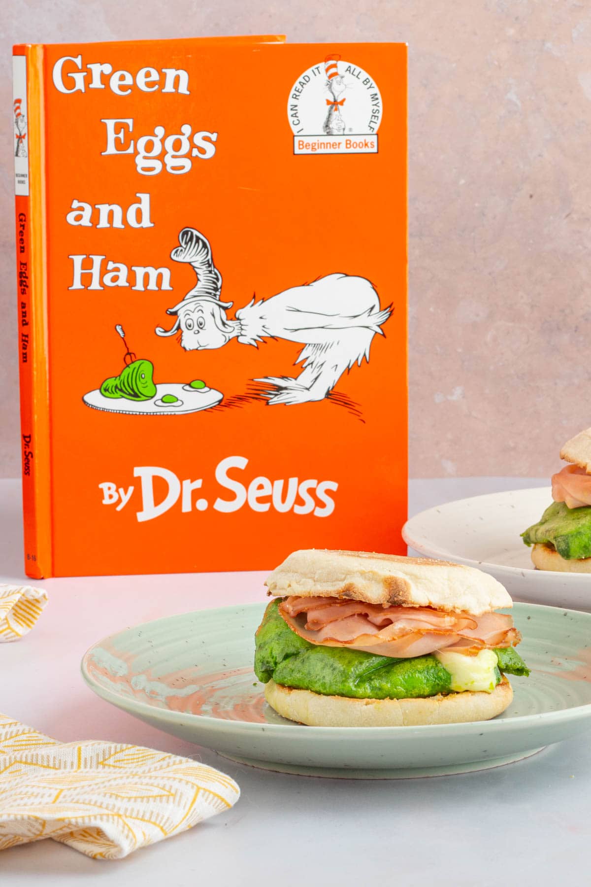 A green eggs and ham breakfast sandwich on a plate next to the book Green Eggs and Ham.