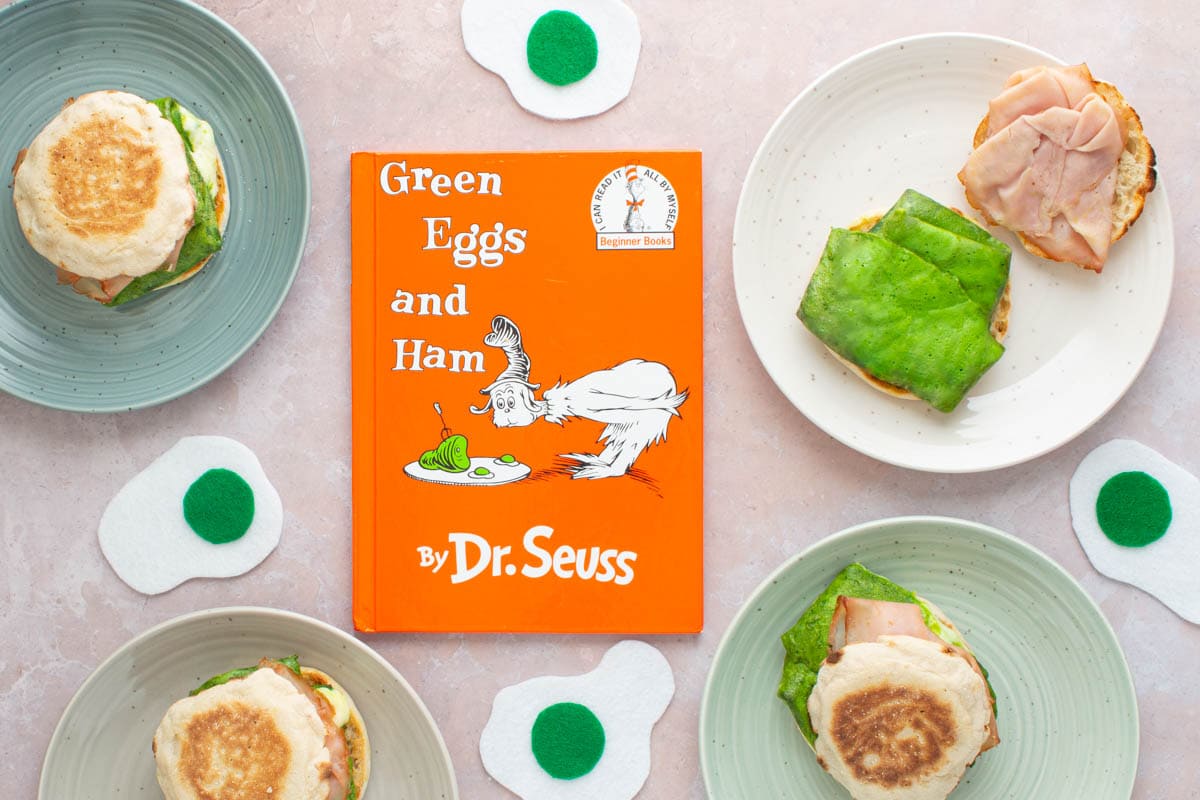 The cover of the book Green Eggs and Ham, with green eggs and ham breakfast sandwiches next to it on plates.