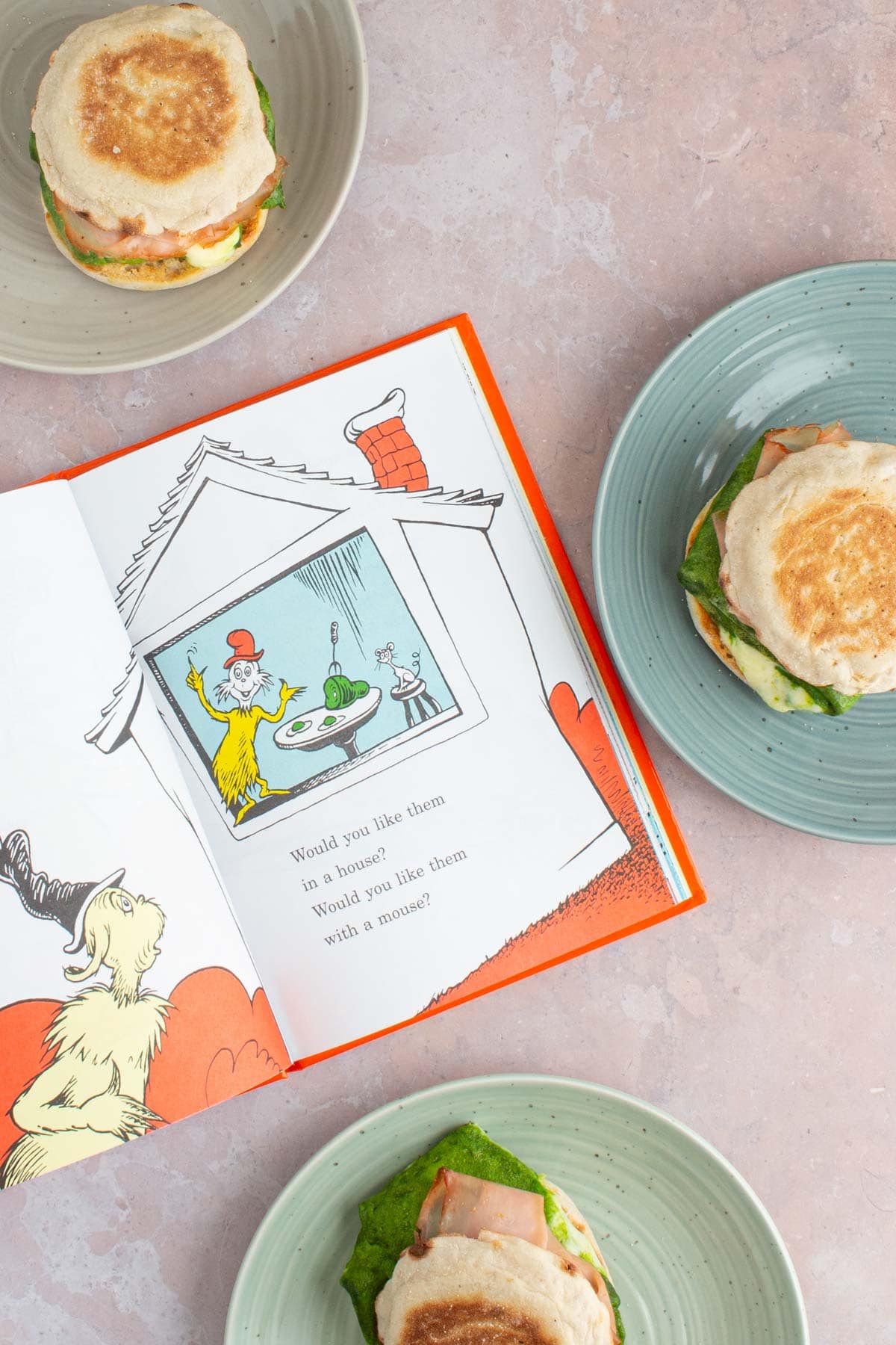 The book Green Eggs and Ham open to a page of illustration, with green eggs and ham breakfast sandwiches on plates next to it.