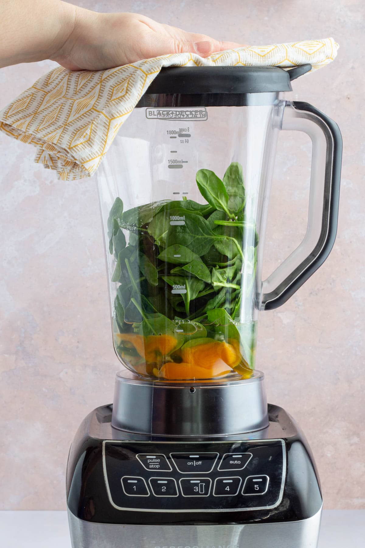 A hand using a kitchen towel to hold the lid of a blender with spinach and eggs in it.