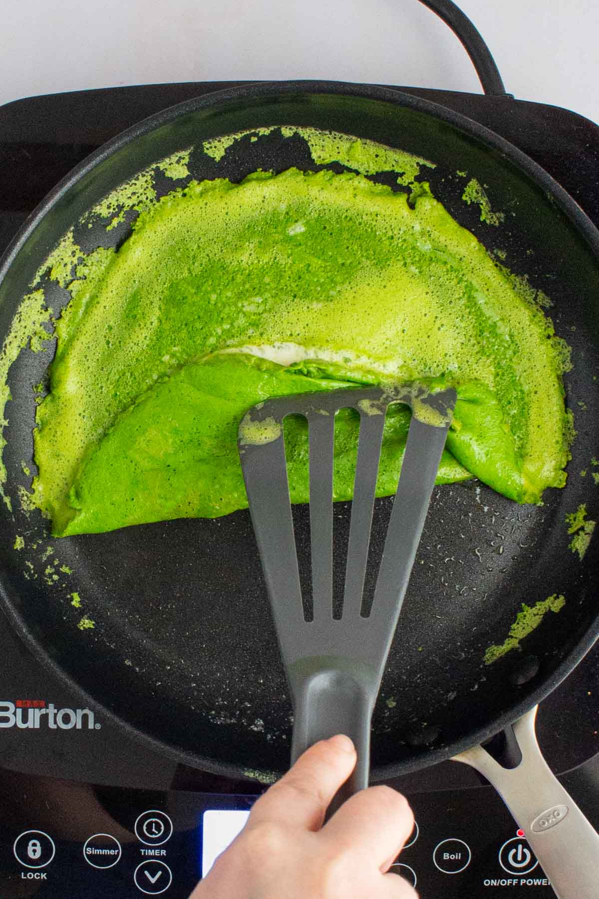 A spatula folding the edge of cooked eggs over cheese in a skillet for green eggs and ham breakfast sandwiches.