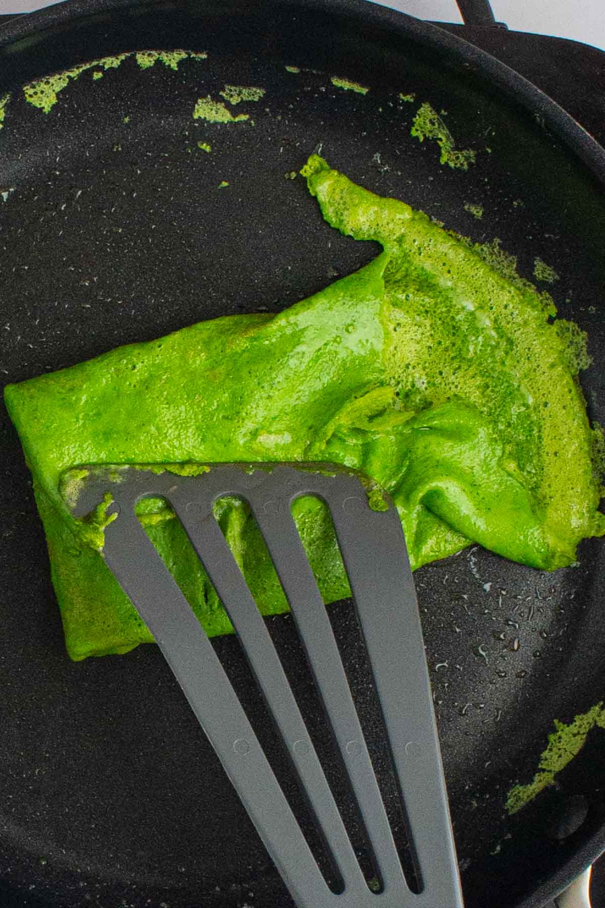 A spatula folding the edge of cooked eggs over cheese in a skillet for green eggs and ham breakfast sandwiches.