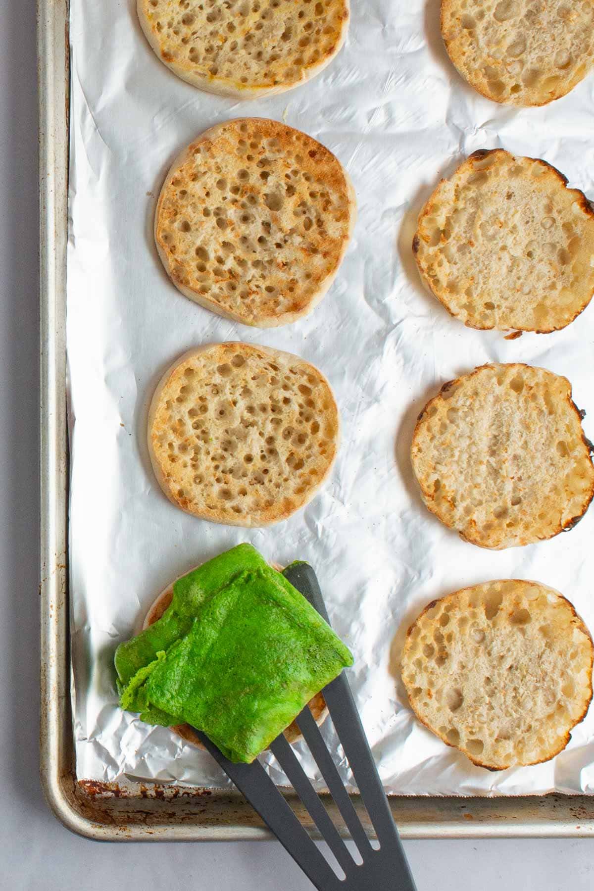 A spatula transferring cooked eggs to an English muffin for green eggs and ham breakfast sandwiches.