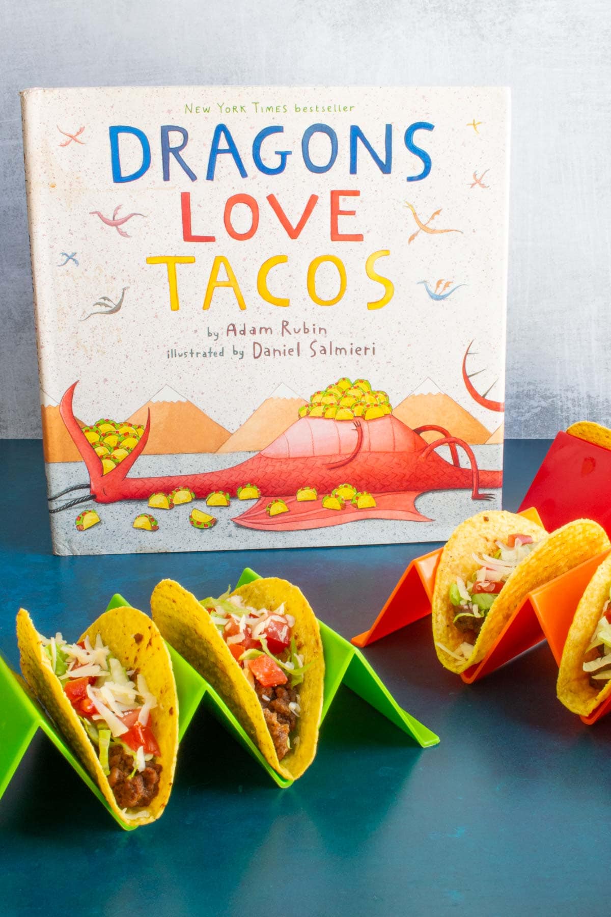 The cover of the book "Dragons Love Tacos" next to taco holders holding assembled Ground Beef Tacos.