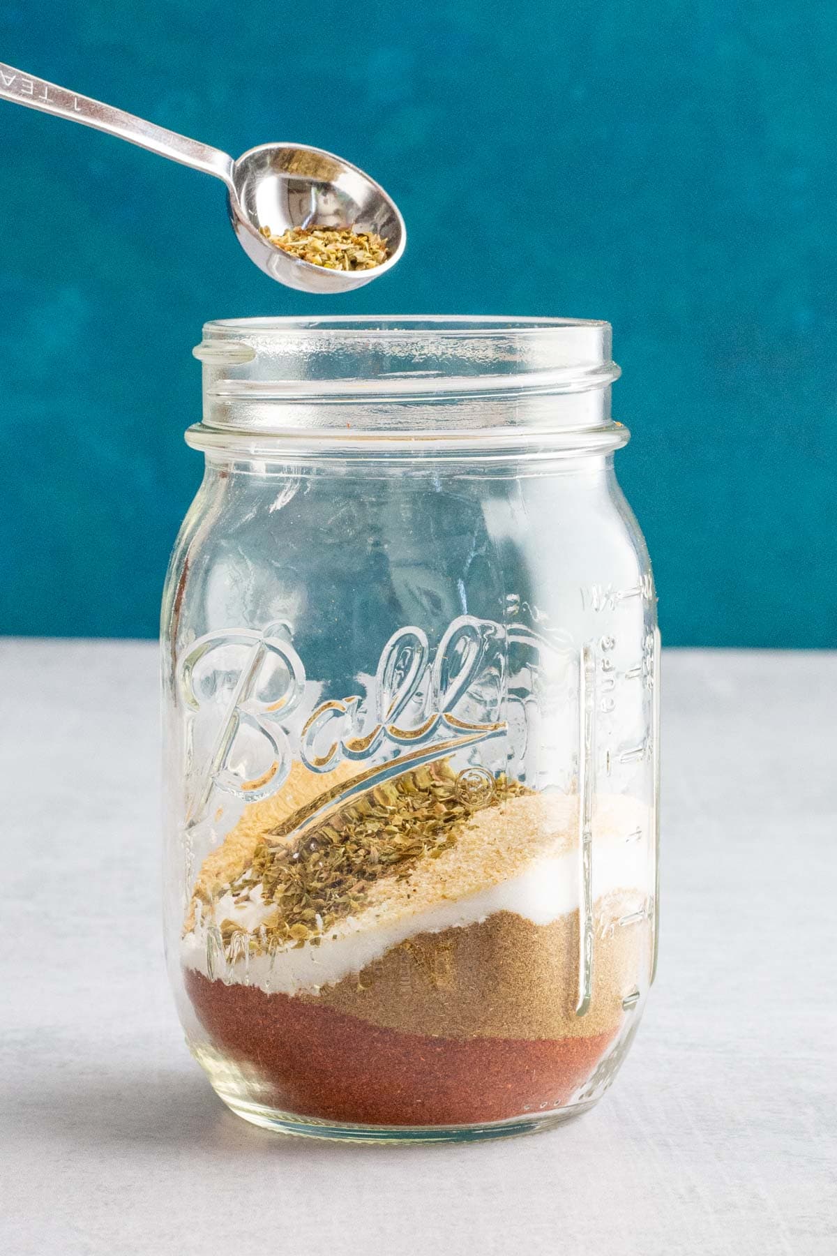 A teaspoon pouring dried oregano into a jar of Homemade Taco Seasoning.