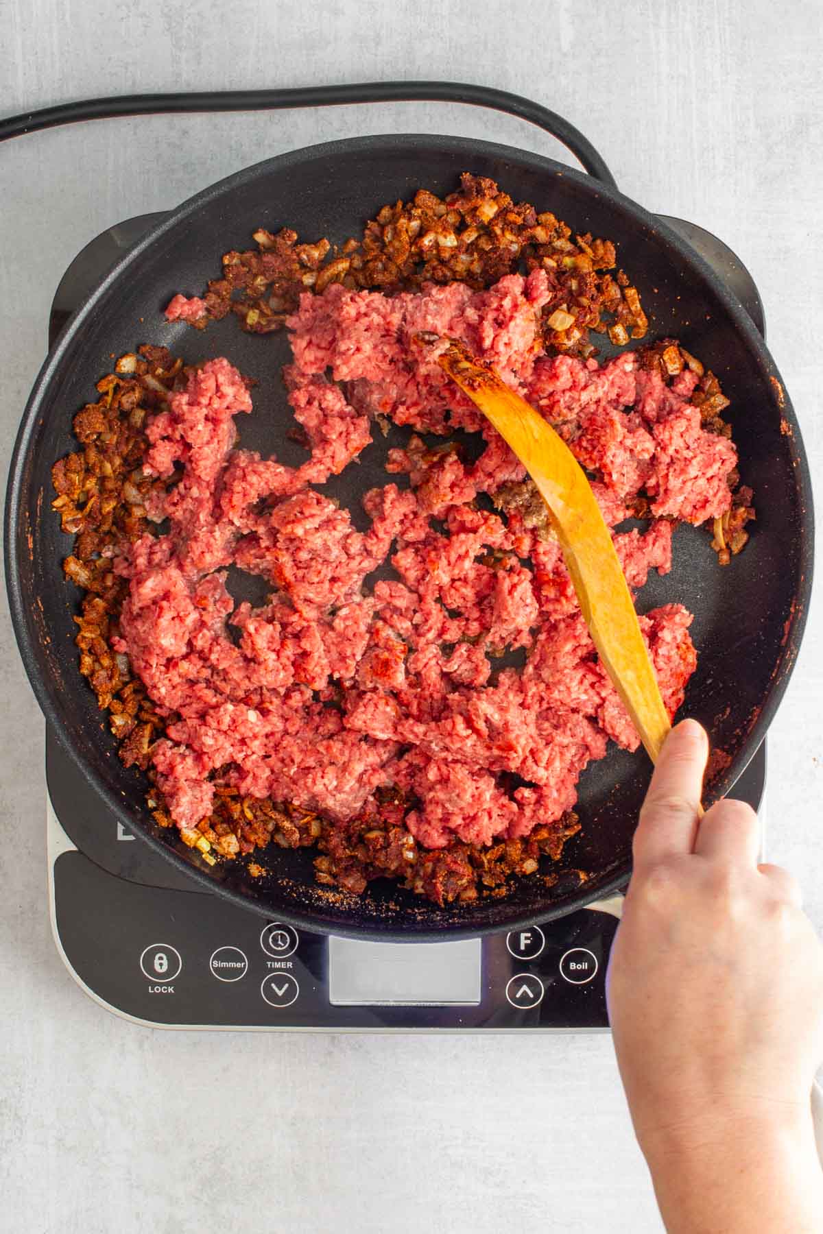 A hand using a wooden spoon to break up ground beef into seasoned onions in a skillet for Ground Beef Tacos.