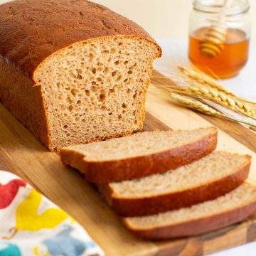 A loaf of Easy Honey Wheat Sandwich Bread cut into slices on a cutting board, with stalks of wheat and a jar of honey nearby.