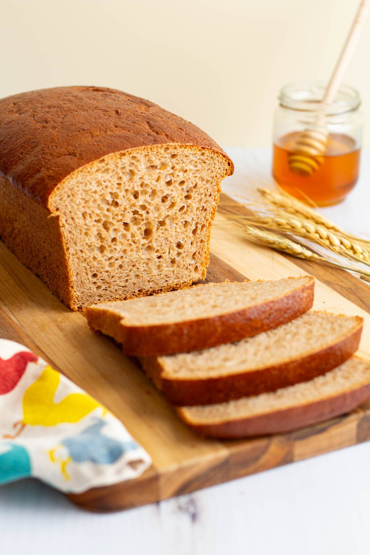 A loaf of Easy Honey Wheat Sandwich Bread cut into slices on a cutting board, with stalks of wheat and a jar of honey nearby.