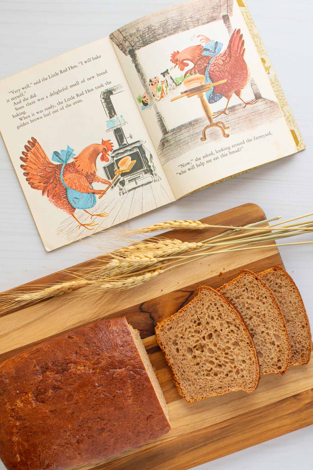 A sliced loaf of Easy Honey Wheat Sandwich Bread on a cutting board next to illustrations from the book The Little Red Hen.