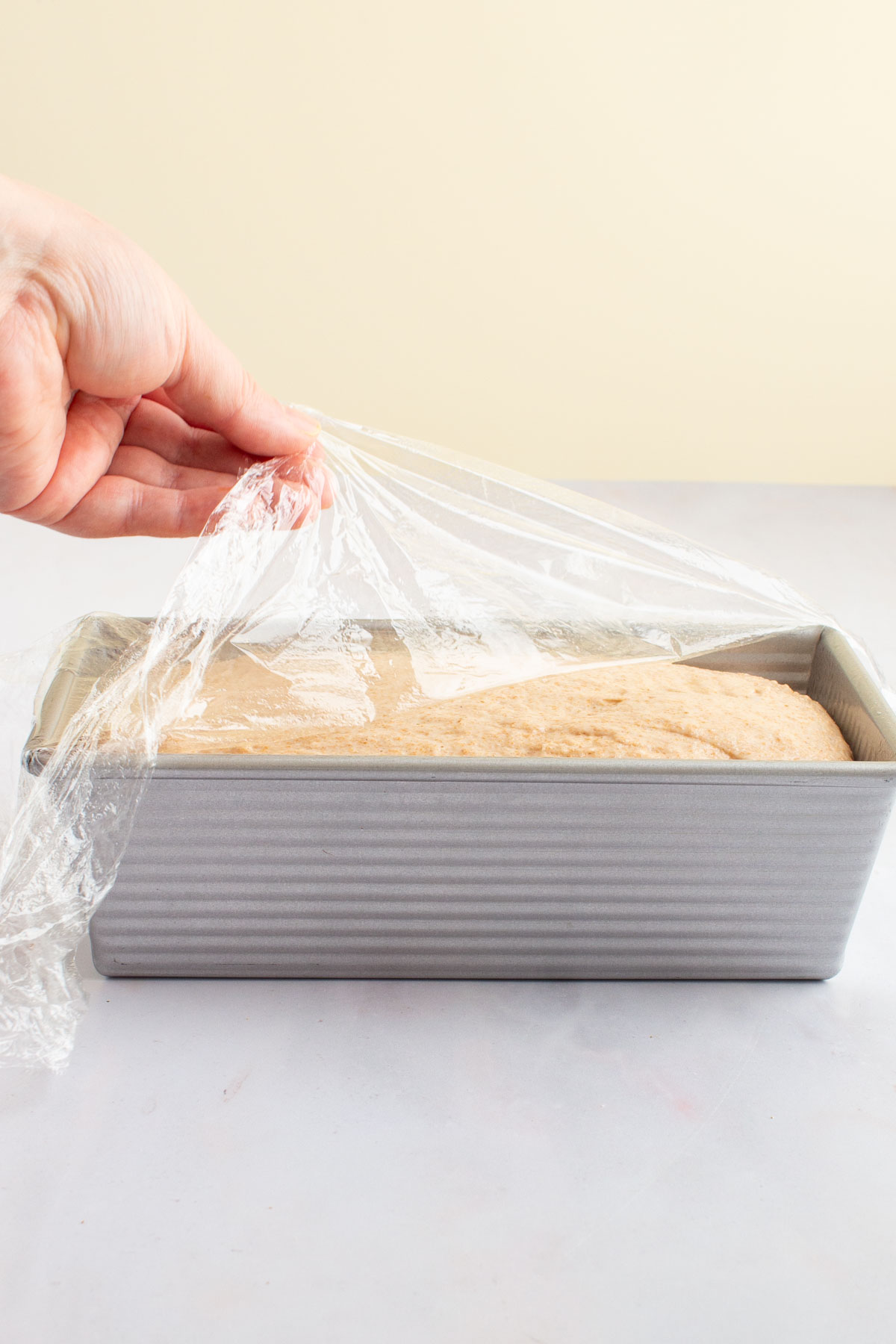 A hand peeling plastic wrap from the surface of partially-risen Easy Honey Wheat Sandwich Bread in a loaf pan.