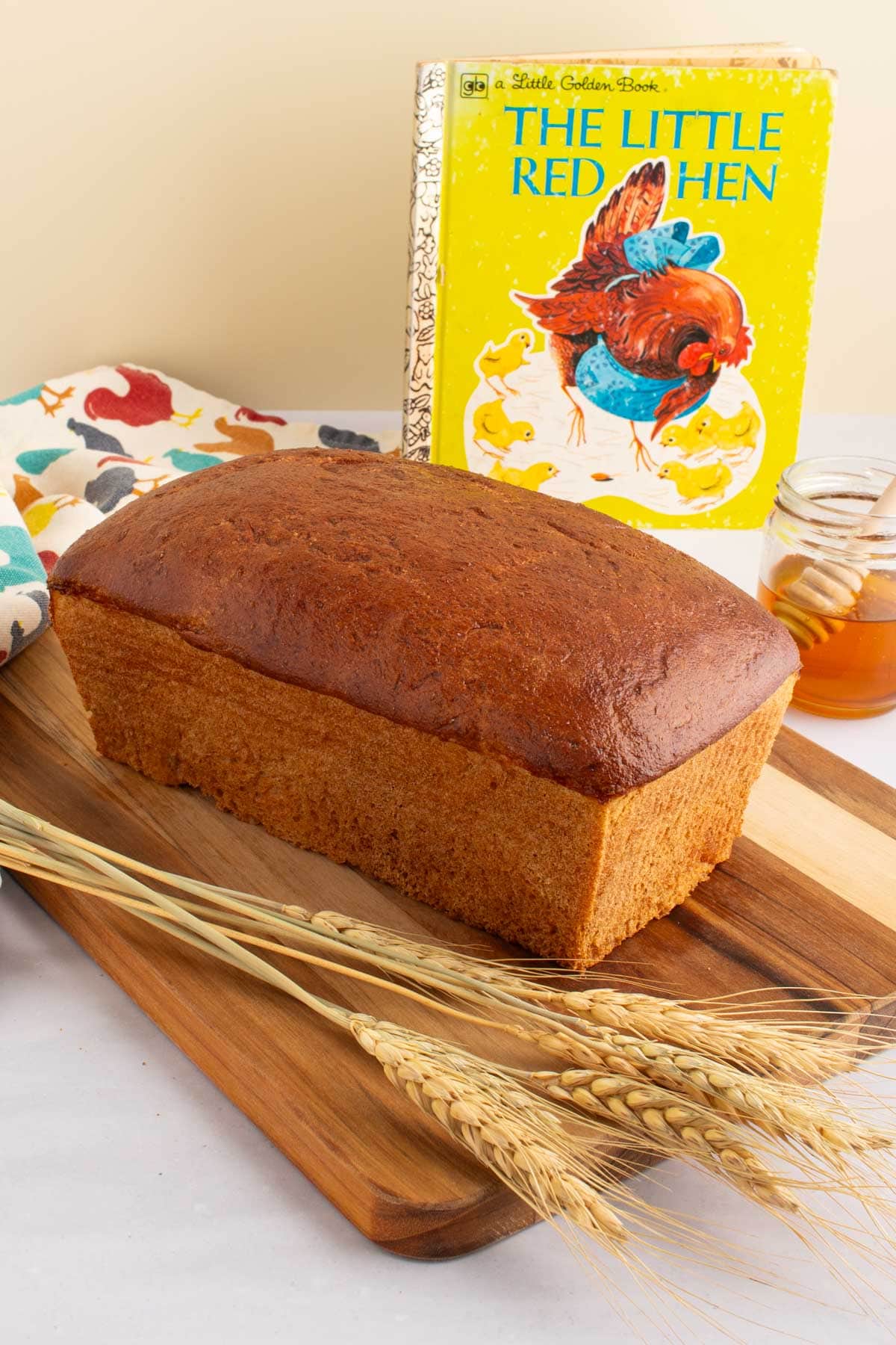 A baked loaf of Easy Honey Wheat Sandwich Bread on a cutting board with the cover of the book The Little Red Hen in the background.