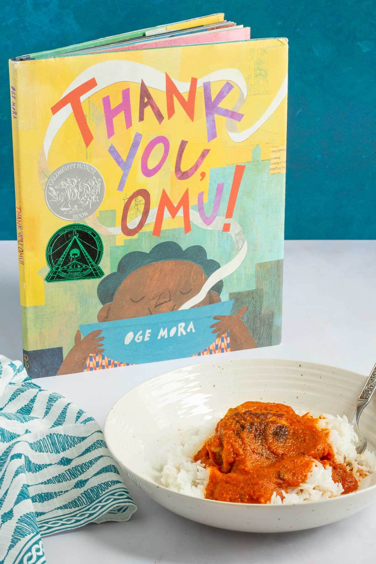 A serving bowl of Nigerian Red Stew over rice with the book Thank You, Omu.